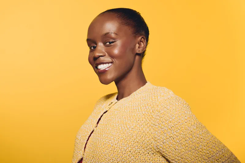 Smiling African woman with dark skin wearing a yellow knitted sweater poses confidently against a bright yellow background. Concept of happiness, fashion, and beauty.