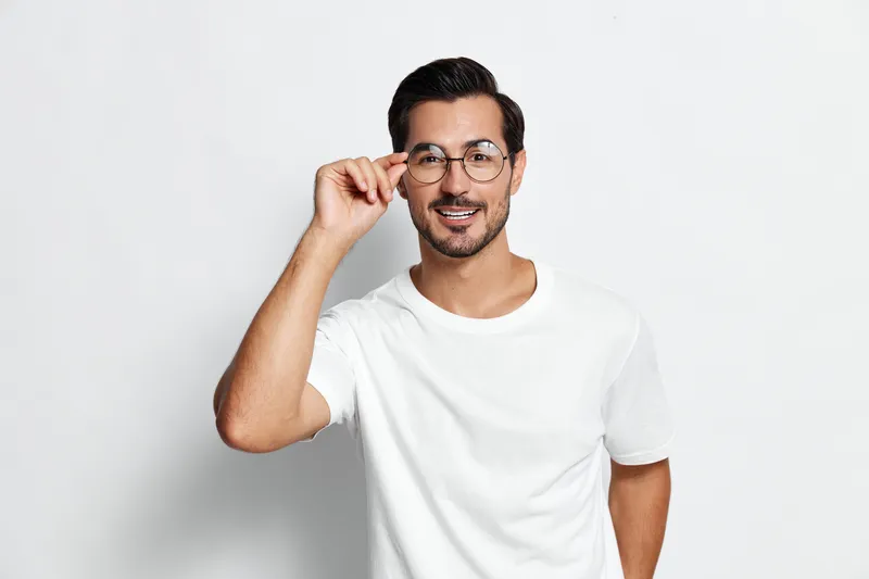 Smiling man adjusts his glasses, wearing a casual white t-shirt while looking confidently at the camera. Isolated on a plain light background studio portrait.