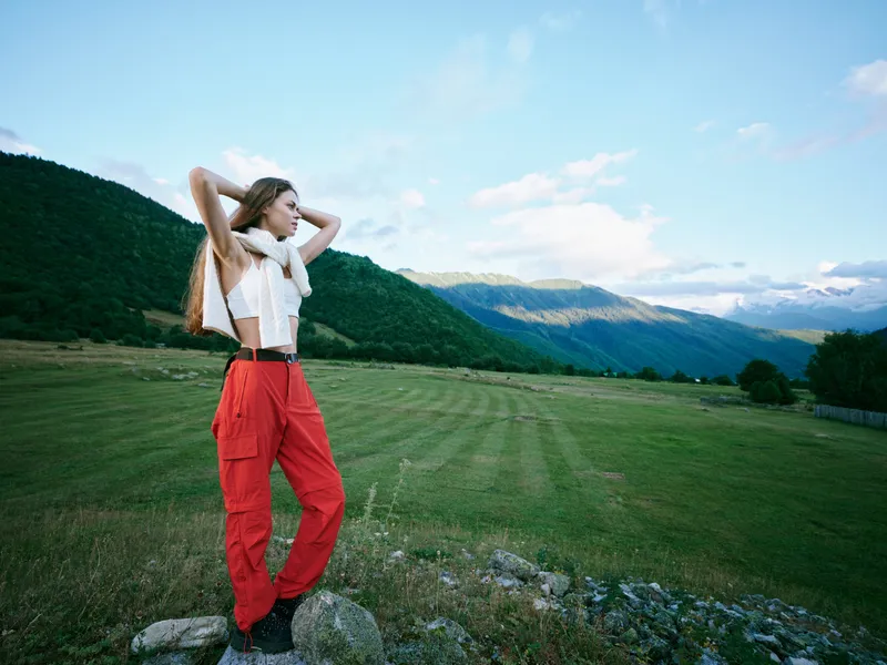 Outdoor landscape field mountains, a woman standing in a grassy plain wearing a white top and red pants, with hands behind head, enjoying expansive space and fresh air under a bright sky.