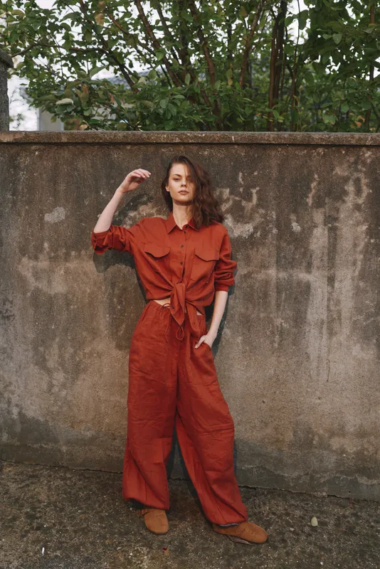Young woman in a rust jumpsuit stands against a weathered wall outdoors, striking a confident pose with one hand in her pocket