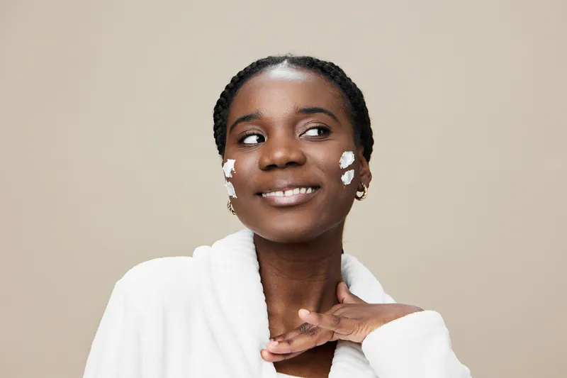 Smiling Black woman applying skincare cream at home, wearing white robe, showcasing healthy skin care routine with a neutral background