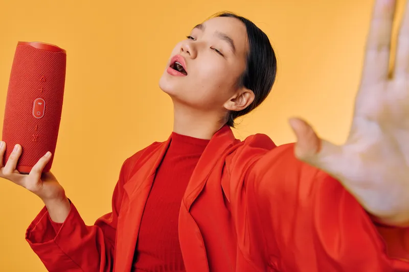 Woman in red jacket and sweater holding portable speaker while singing with closed eyes and hand raised against orange background. Indoor, technology, music and enjoyment with device.
