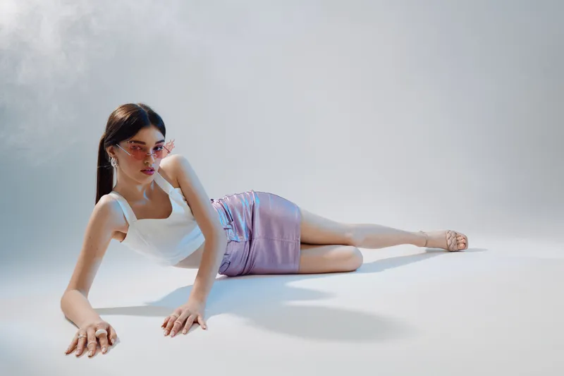 Young woman in a white top and purple skirt lies on the floor wearing pink sunglasses. Studio shot with soft lighting. Concept of woman beauty fashion and modern style.