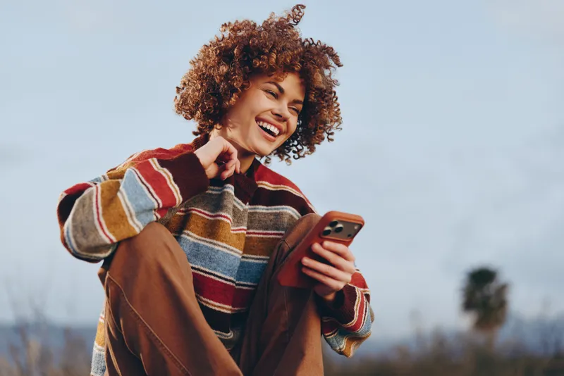 Smiling woman lifestyle outdoors in winter wearing striped sweater and coat, curly hair framing face, holding smartphone and chatting, casual day mood, natural light, blurred background