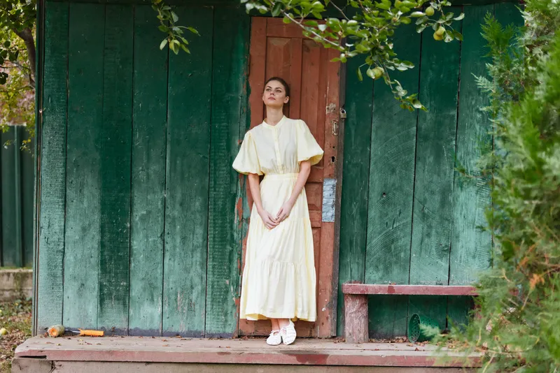 A woman in a pale dress stands on a wooden porch beside a green weathered wall, posing with calm elegance and soft light that highlights the serene outdoor mood.
