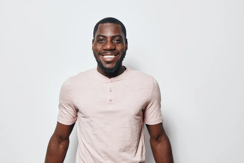 Smiling African American man with dark skin wearing light pink t shirt standing against white background. Portrait of happy black male with short hair and beard looking at camera.