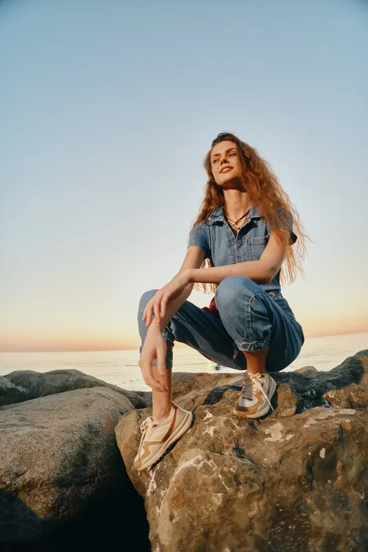 lifestyle woman in modern boho-western denim street style crouching on rocks at sunset with relaxed expression and film color tones