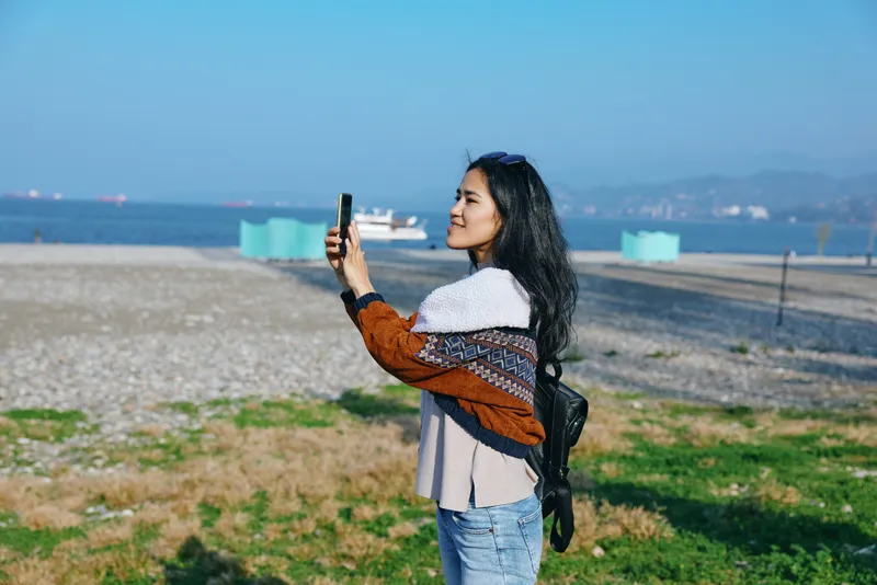 Selfie, woman on a sunny beach taking photo with smartphone, backpack, casual outfit, sea horizon in background, travel mood and outdoor photography