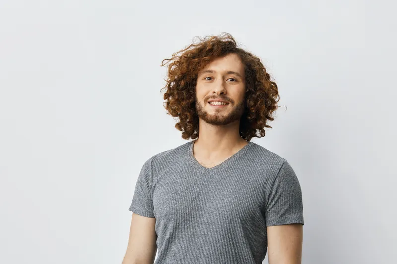 Happy young man with curly hair wearing a gray t shirt, smiling brightly against a light background, showcasing confidence and positivity in a modern setting
