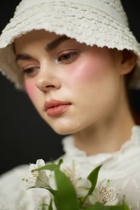 Delicate portrait of a model in a white hat, showcasing soft makeup and holding flowers, evoking a serene and natural aesthetic