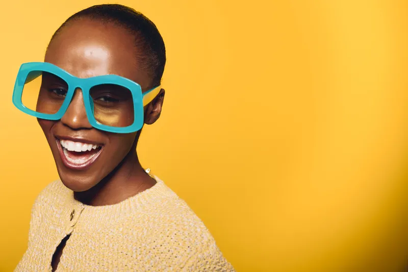 Happy dark skinned African woman wearing large blue glasses smiles widely against yellow background. Concept of joy, fashion, and vibrant lifestyle with female model in studio lighting.