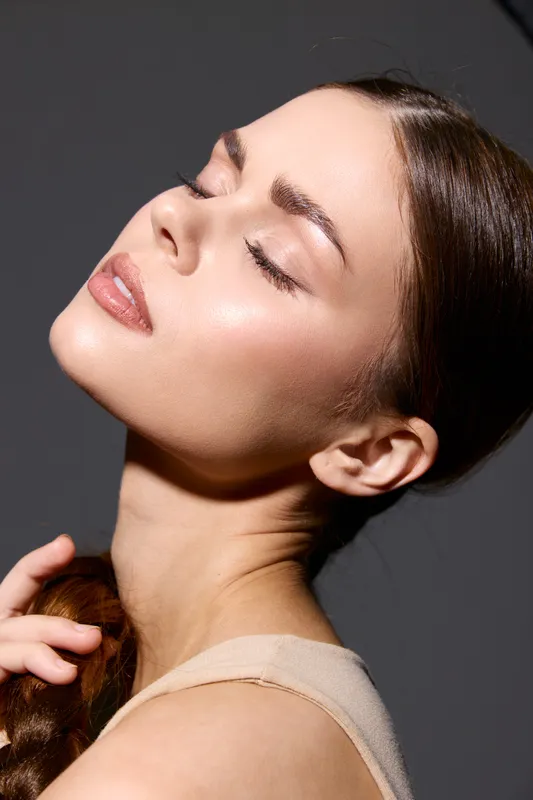 beauty woman with glowing skin and closed eyes poses gracefully against dark background, showcasing natural makeup and smooth complexion in soft lighting.