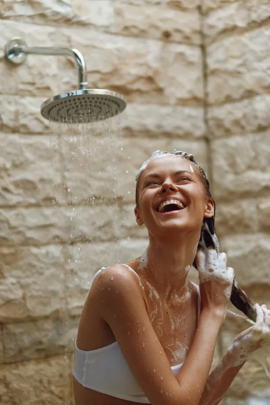 smiling woman washing hair under rain shower with soap foam on skin, joyful expression, natural stone wall background, wellness and relaxation concept