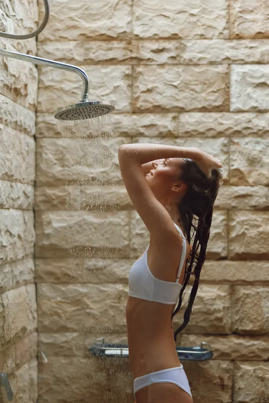 Woman showering outdoors under rainfall with stone wall background, wearing white underwear, enjoying a refreshing natural bath in warm daylight.