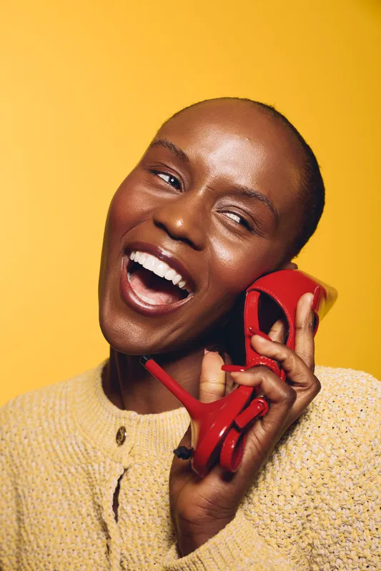 Happy black woman with dark skin laughs holding red high heel shoe near face on yellow background. Concept for fashion, beauty, joy, and lifestyle with African ethnicity model.