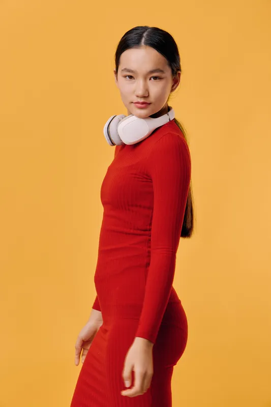 Young woman wearing red dress and white headphones posing against yellow background, looking at camera with confident expression and long dark hair tied back in ponytail.