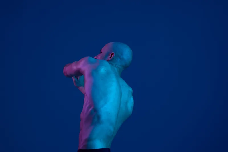 Muscular man posing in dramatic lighting, showcasing strong physique and artistic shadow play. The vibrant color contrasts enhance the visual impact, creating an engaging aesthetic.
