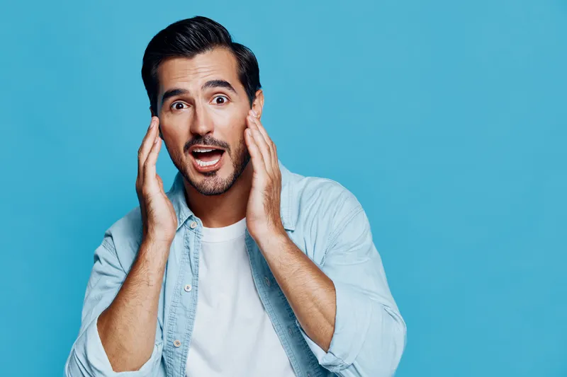 Surprised happy young man with short black hair wearing light blue denim jacket, holding his face in astonishment against solid blue background. Emotions concept