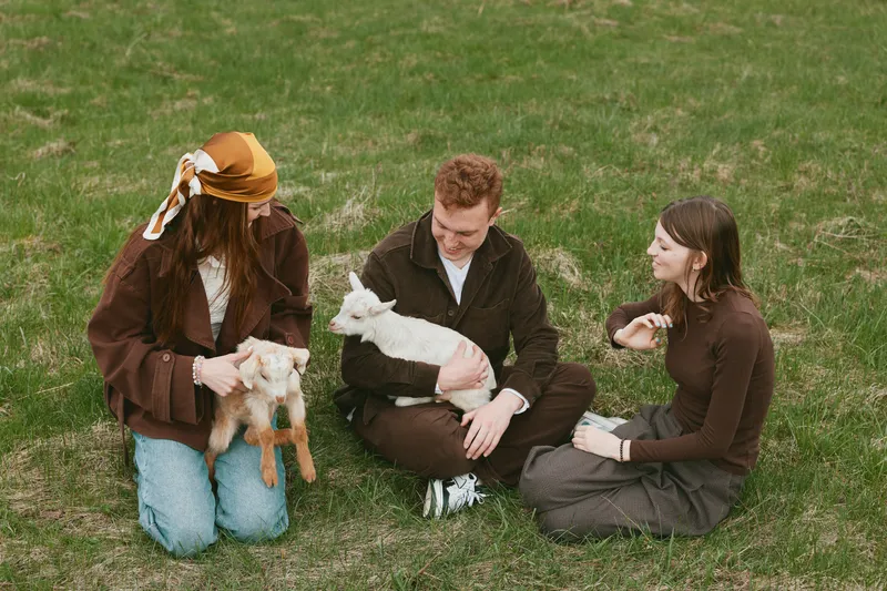 Three people sit on a grassy field with a goat kid, a man cuddles a baby goat, a woman in a mustard hat smiles, another woman listens, creating a warm lifestyle moment of outdoor bonding