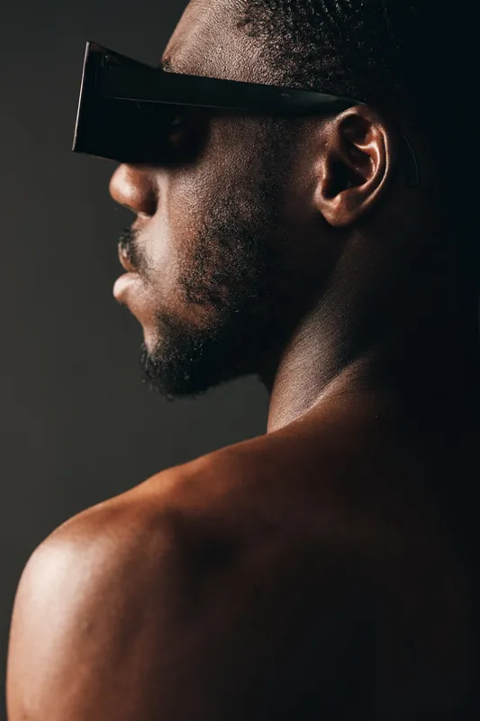 Black man with dark skin wearing futuristic virtual reality glasses in creative conceptual visual style. Side profile, bare shoulders, technology and innovation in digital experience.