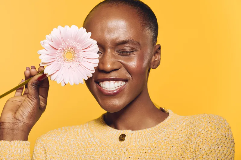 Happy African woman with dark skin closes eyes and smiles holding pink flower near face. Bright yellow background and cozy sweater create warm, joyful mood for beauty and wellness concept.