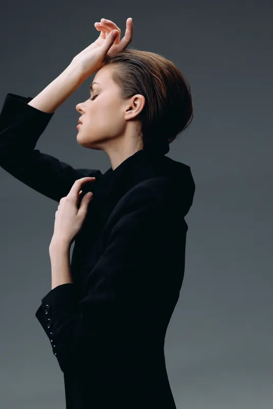 Elegant woman in black jacket posing with eyes closed and hand on forehead against dark background