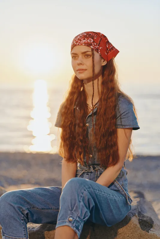 lifestyle woman with long red hair wearing modern boho-western denim street style outfit and red bandana by the beach at sunset, film color warm tones
