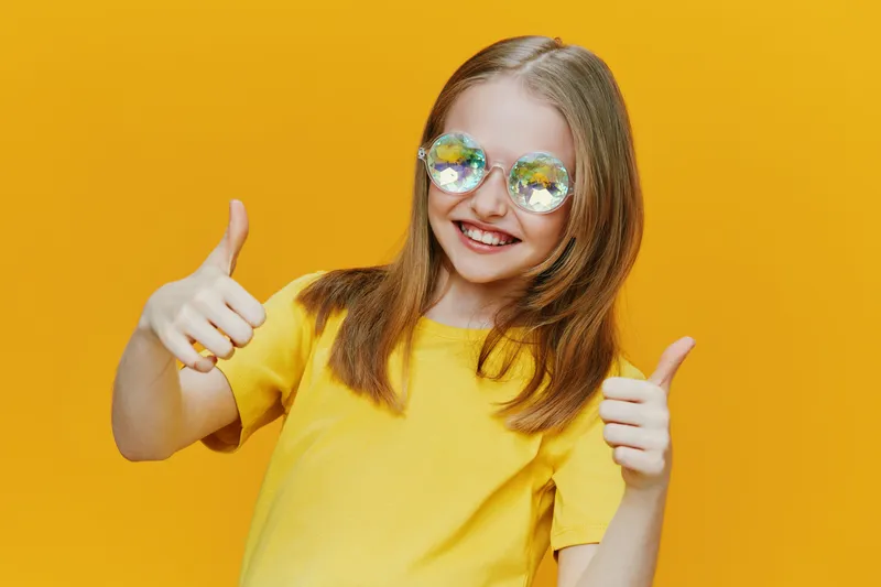 Cheerful girl with sunglasses giving thumbs up against a bright yellow background, expressing happiness and positivity, perfect for summer themed projects