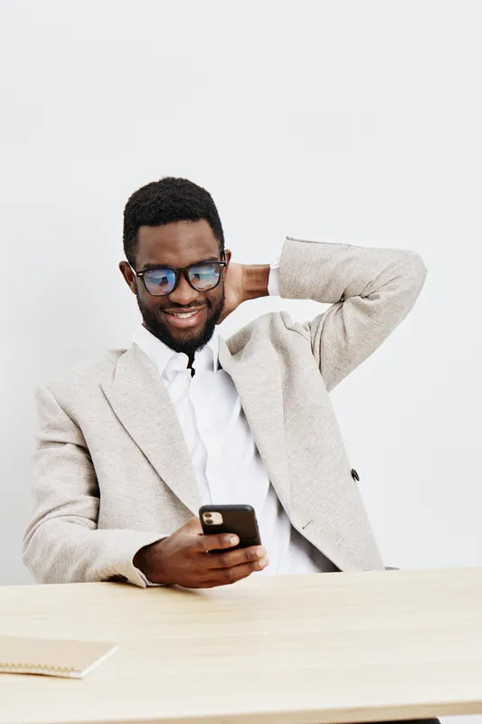 Smiling man with glasses using a smartphone, wearing a beige blazer and white shirt, sitting at a light wooden table in a bright studio, lifestyle concept, casual, cheerful, modern office attire, male