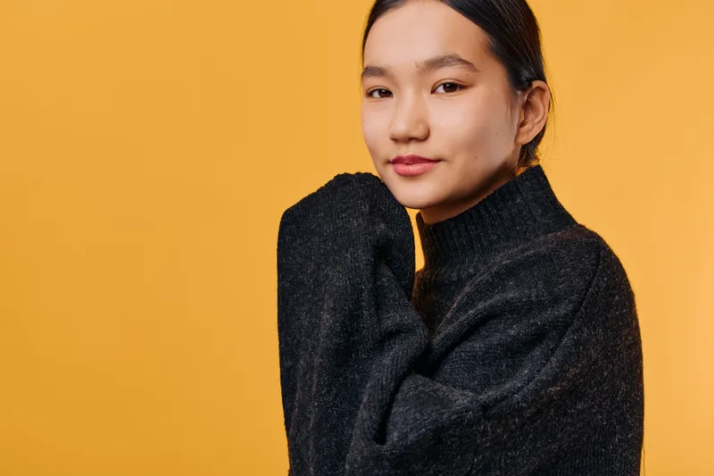 Portrait of young woman smiling and wearing black sweater against orange background. Female model with dark hair looking confident and relaxed in studio lighting.