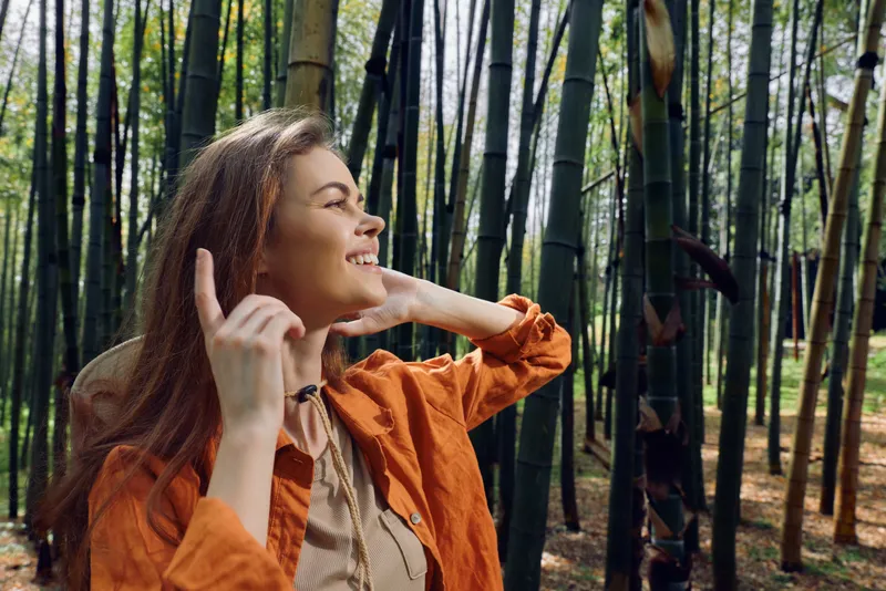 Woman portrait in bamboo forest smiling, nature outdoors scene with happy young traveler in orange jacket enjoying sunshine and peaceful green grove atmosphere.