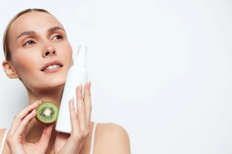 Young woman holds skincare spray bottle and fresh kiwi fruit near face with clear skin and natural makeup isolated on white background with copy space for beauty concept.