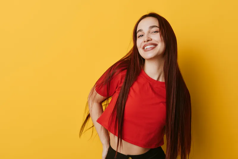 Young woman with long hair smiling and posing in red crop top against yellow background. Casual style, happy expression and confident posture for lifestyle and fashion.