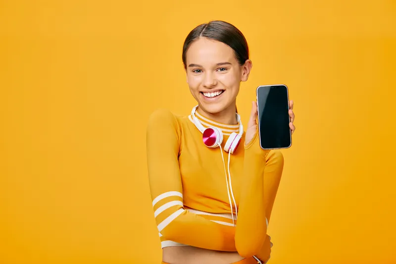 Woman in an orange athletic top with headphones, smiling and holding a smartphone against a vibrant yellow background, illustrating fitness, sport, and digital motivation for wellness campaigns.