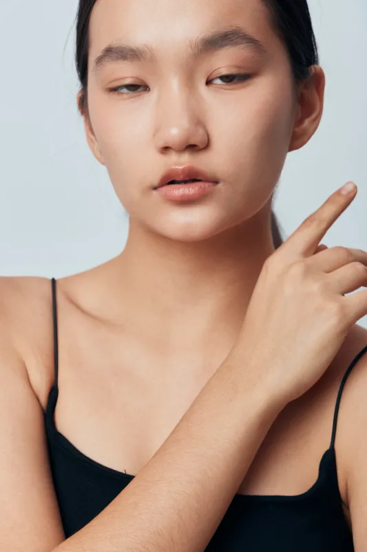 Young woman with smooth skin in black spaghetti strap top posing on light background, closeup portrait with natural makeup and calm expression for beauty and fashion concept.