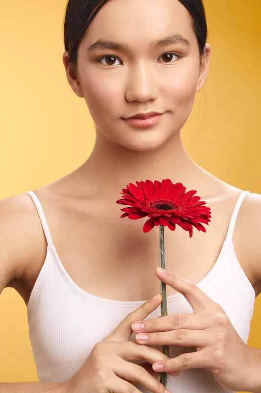 Asian woman presents a serene smile as she holds a bright red flower against a warm backdrop, conveying calm elegance and gentle charm in a studio portrait.