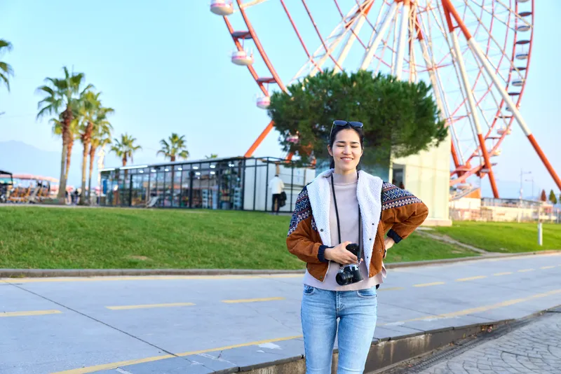 Woman with camera at outdoor amusement park near ferris wheel, casual fashion, smiling stance, travel photography vibe, sunny day, scenic boardwalk setting, lively urban leisure atmosphere