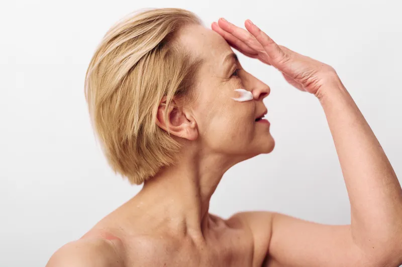 Mature woman with short blonde hair applies white face cream on cheek against white background. Concept of skincare, aging care, wellness, and natural beauty with soft natural lighting.