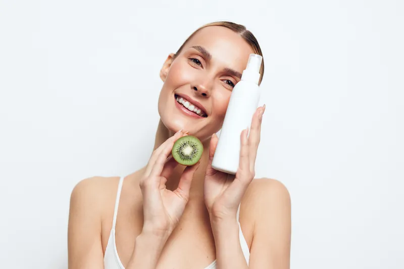 Smiling woman holds white cosmetic bottle and fresh kiwi fruit near face isolated on white background showing skincare and natural beauty concept with healthy lifestyle.