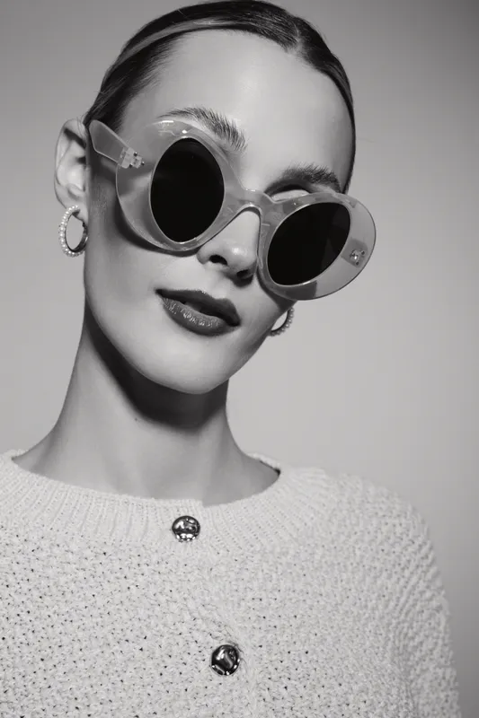 Slim Caucasian woman with pale skin wears round transparent sunglasses and hoop earrings, dressed in textured sweater. Studio portrait in black and white with fashion and style concept.