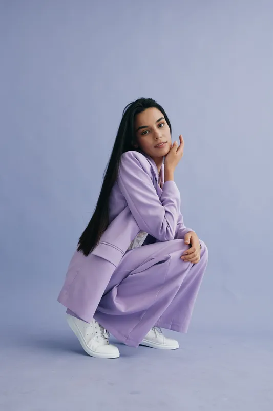 Young woman wearing a pastel purple suit poses against a soft lavender background, showcasing modern fashion with a calm and confident expression in studio lighting.