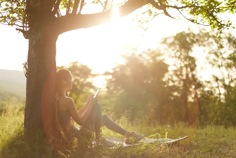 Young woman enjoying a peaceful moment reading under a tree in the park at sunset, surrounded by nature and warm light, evoking relaxation and tranquility