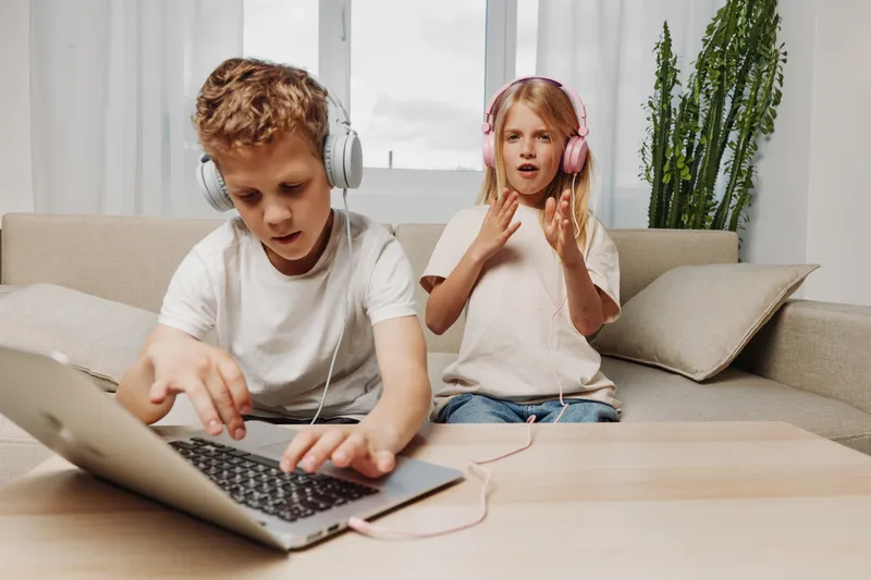 Two children with headphones use laptop at home. Boy types on keyboard while girl sings with hands raised. Casual clothes, cozy interior, natural light. Concept for technology and creativity.