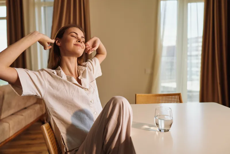 Woman morning stretching and relaxation at home in sunlight, sitting at table with glass of water, casual loungewear and peaceful wellbeing, enjoying quiet slow routine.