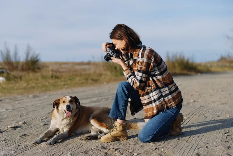 Photographer kneeling in outdoor sandy landscape with a camera, capturing a loyal dog along a quiet trail, showcasing nature, leisure, and companionship under a bright sky