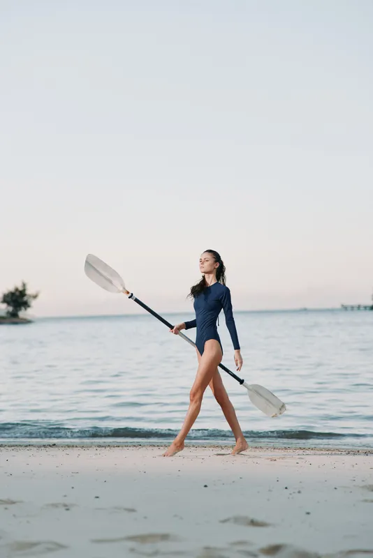 A lone figure walks along a wide sandy beach, carrying two paddles above the water, with a calm sea and clear sky creating a peaceful, minimalist coastal scene.