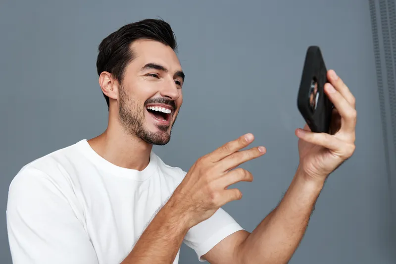 Happy smiling man wearing a white t-shirt looking at his smartphone, engaging in a joyful video call, isolated on a soft gray background. Modern communication concept.