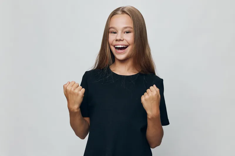Happy girl with long hair wearing black t shirt smiling and showing fists in victory pose on grey background, cheerful child celebrating success or achievement indoors.