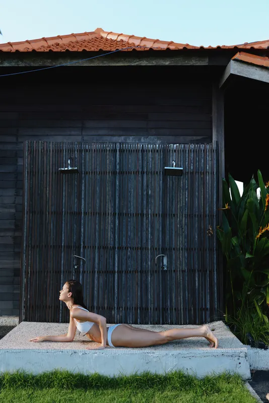 Young woman practices yoga outdoors in cobra pose wearing white sportswear near wooden wall with tropical plants in background during sunny day.