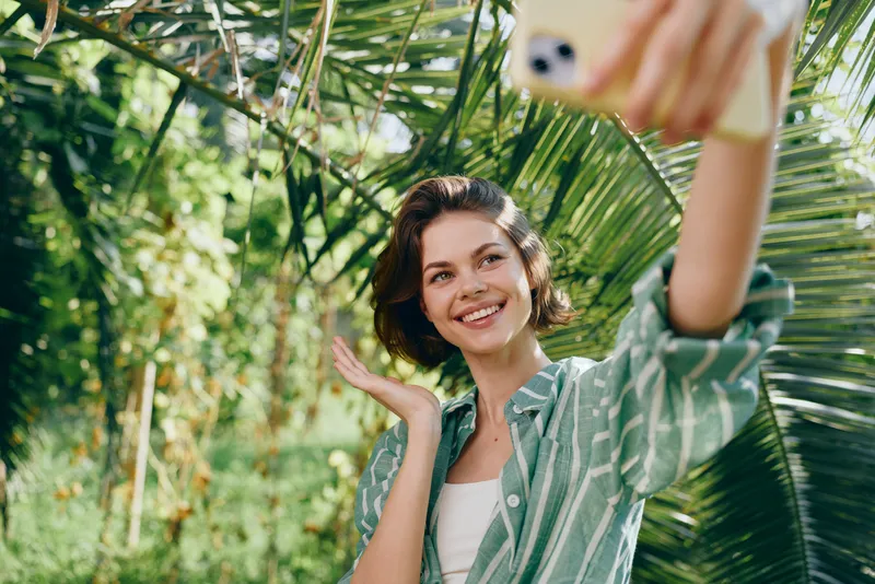 Young woman takes a bright selfie outdoors among lush tropical leaves, wearing a striped shirt and a cheerful expression.